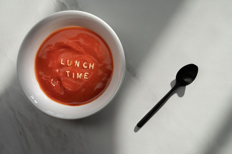 A flat lay image of tomato soup with 'Lunch Time' spelled out, accompanied by a black spoon on a marble surface.
