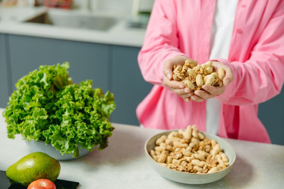 Hands presenting peanuts with a bowl, lettuce, avocado, and tomato nearby on kitchen count