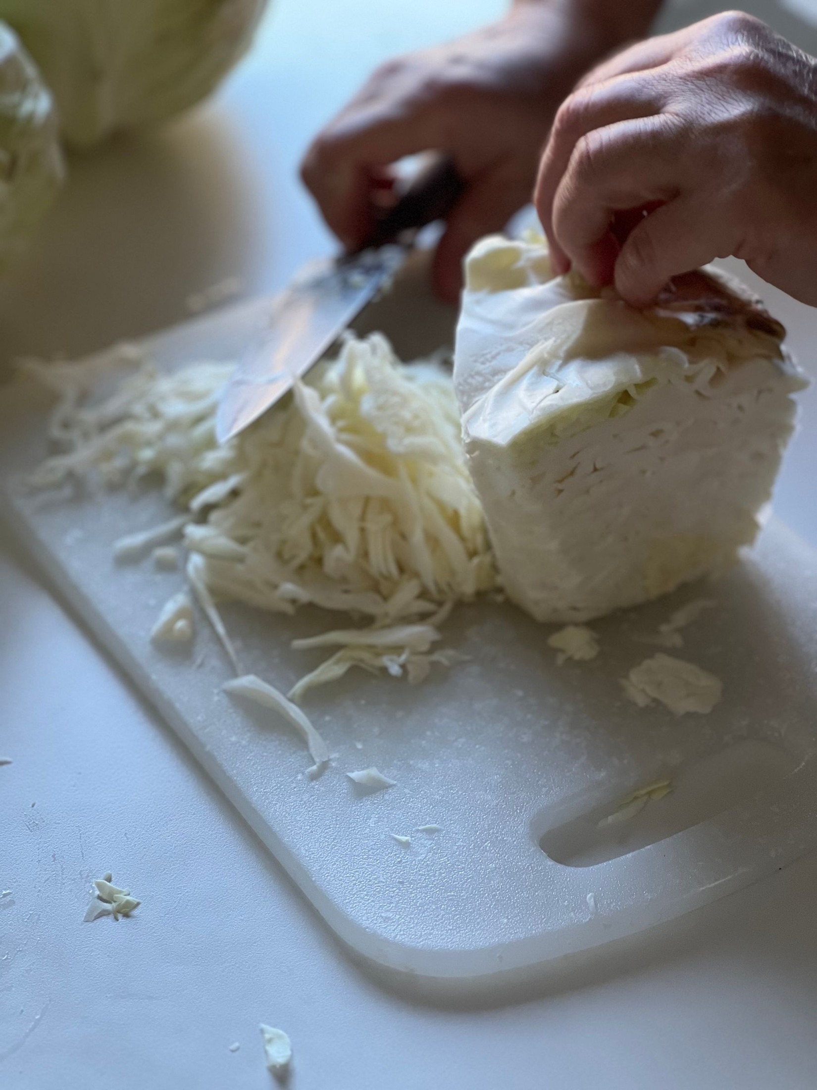 Hands slicing a white-cabbage wedge into shreds on a pale cutting board, with loose shredded cabbage on the neutral kitchen work surface.