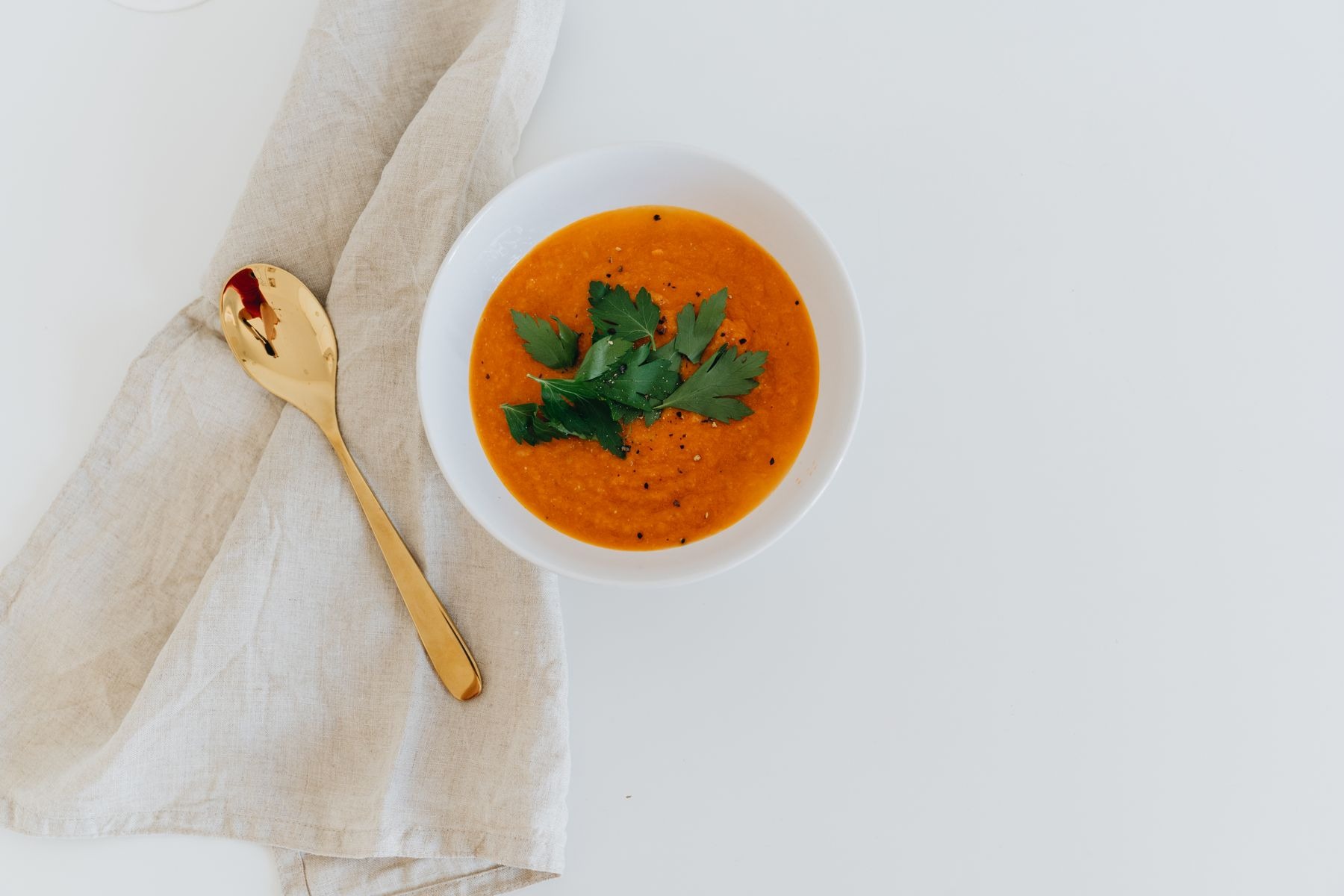 Carrot soup garnished with herbs beside a gold-colored spoon on white.
