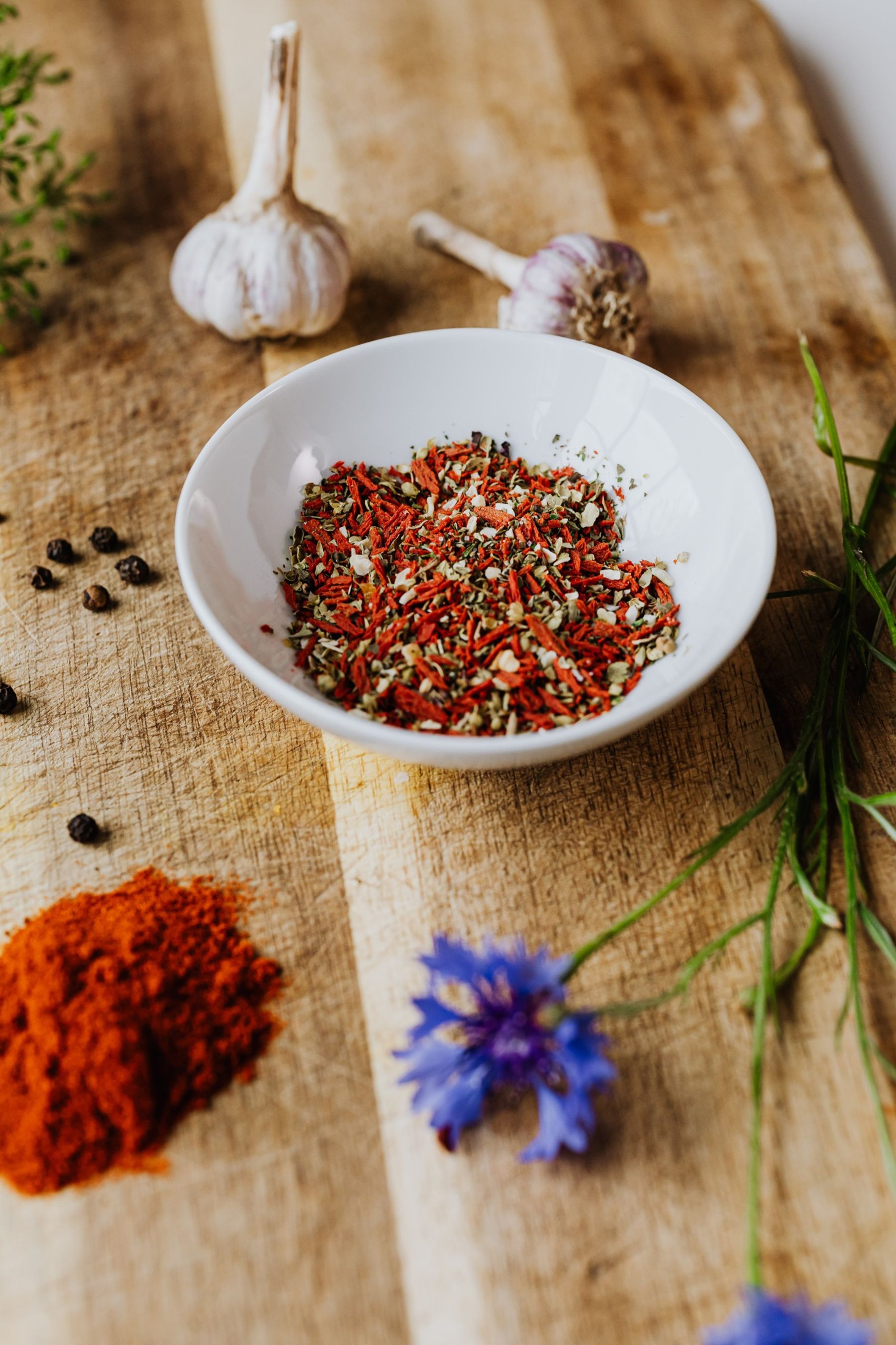 Mixed herbs and seasonings in a ceramic bowl with garlic and peppercorns nearby.