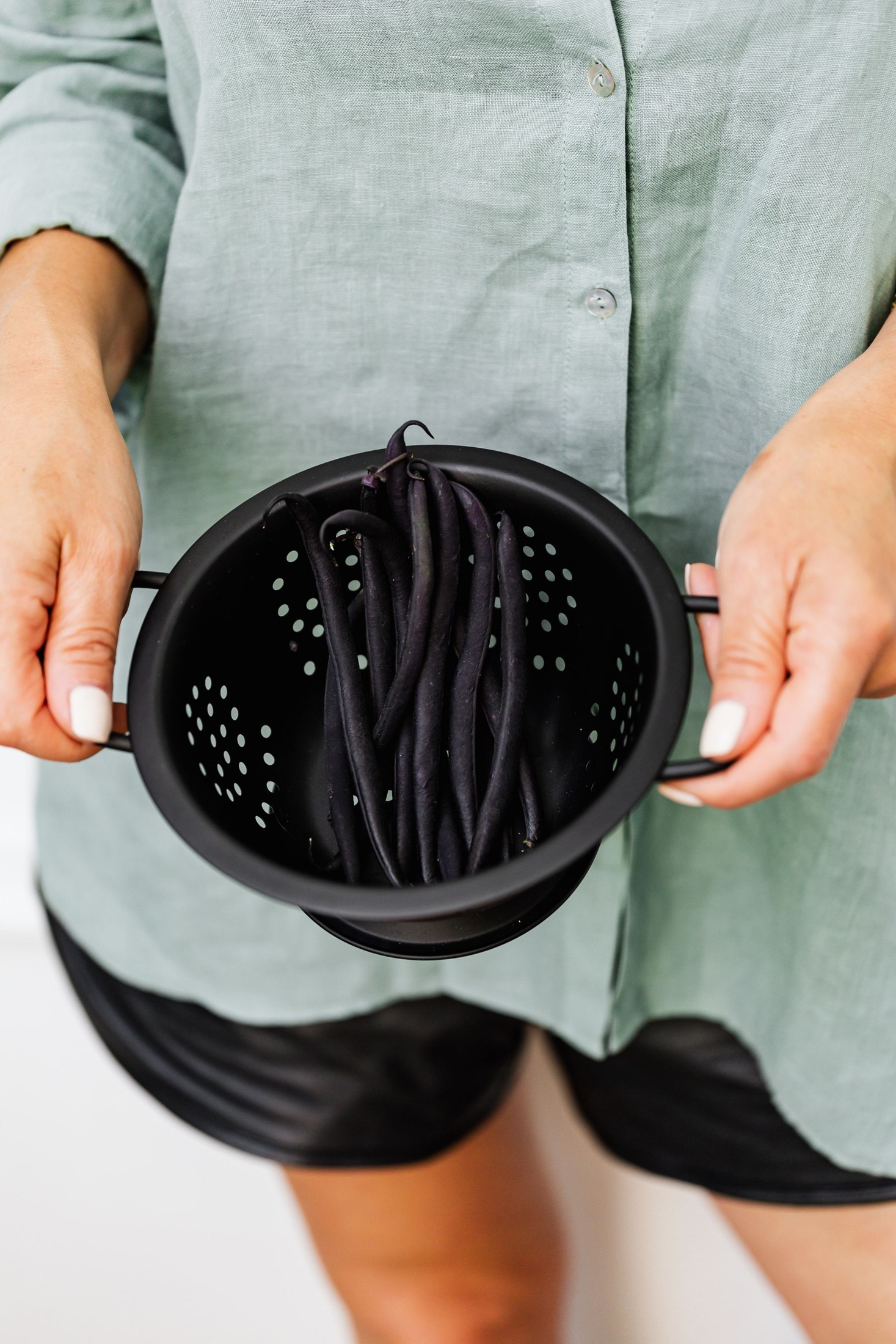 Person holding a black colander of long dark-purple bean pods; these are NOT shelled black beans.