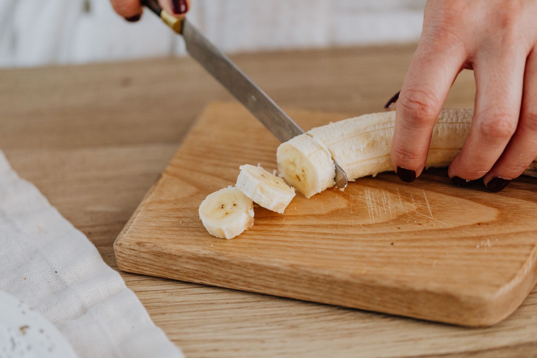 Hands slicing a peeled banana on a wooden board.