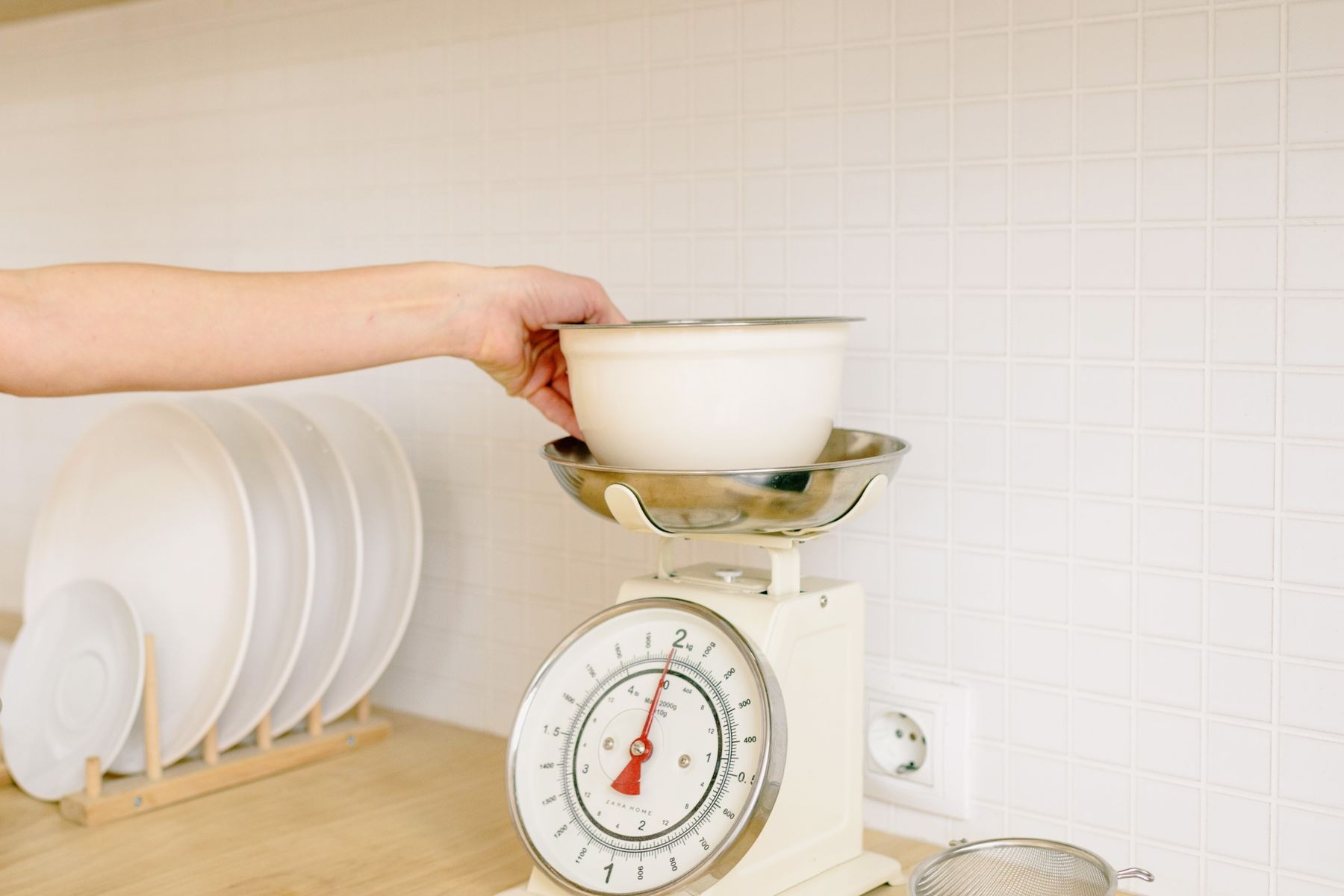 Hand steadying an ingredient bowl on a pale green analog kitchen scale; eggs nearby.