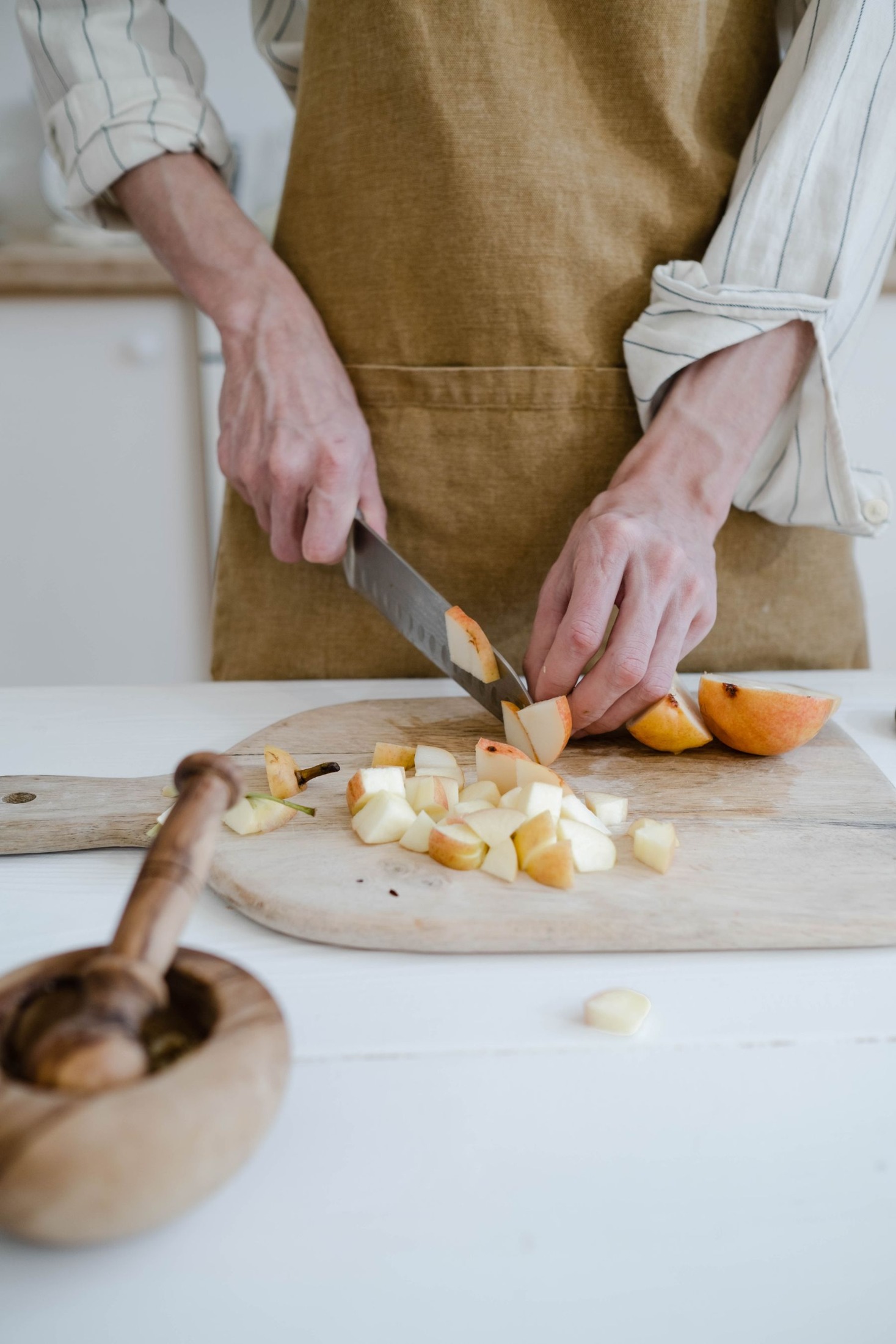 Hands cutting apple pieces on a wooden chopping board at a white kitchen worktop; neutral apron and pale background.