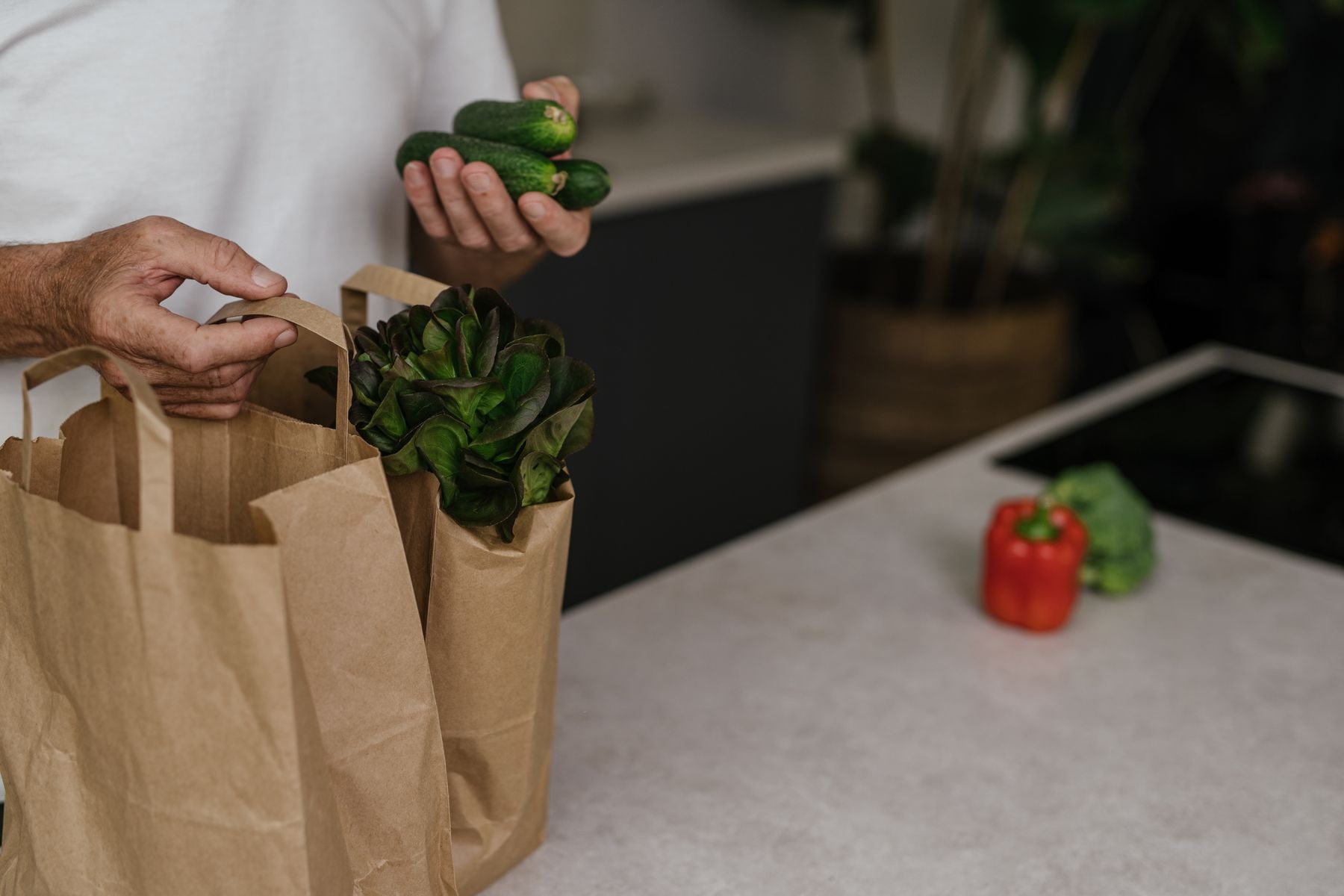 Fresh vegetables being placed in a paper grocery bag at a kitchen counter.